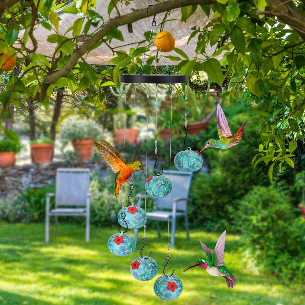 Hummingbird feeder hanging in a garden with greenery and chairs in the background.